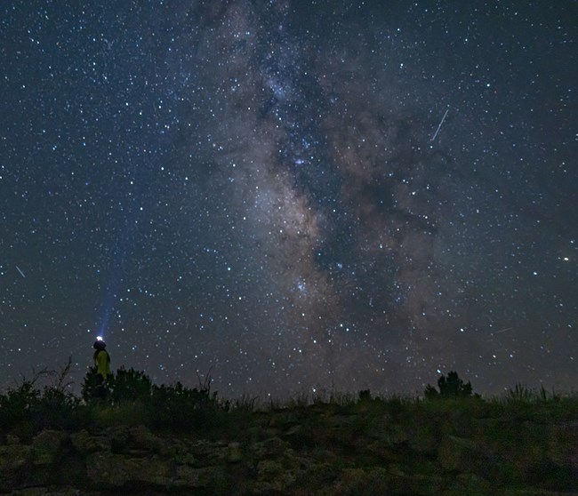 A hiker with a headlamp looks up at the star-filled night sky.  The Milky Way Galaxy creates a band of light blue and silver in the sky.