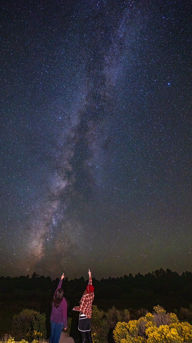 Two hikers point to the night sky, each using an extended arm and their pointer finger.  A silvery-blue band of stars stretches from the horizon up into the sky.