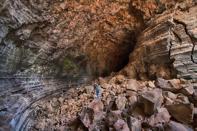 Two cavers are dwarfed by the walls and rubble within a sunlit cave.