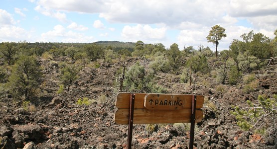 A wooden sign with an upward-facing arrow reads "Parking."  It is the only man-made feature on a rugged lava flow dotted with bushes.