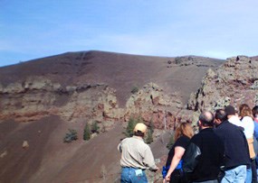 A group of hikers huddle together as they look away and down into an out-of-frame volcanic crater.  Rock cinders drape over a thick layer of rock that stretches across from left to right.