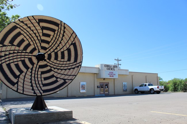 A repurposed satellite dish art piece stands in front of a single-level building with a marquee reading "Cibola Arts Council."