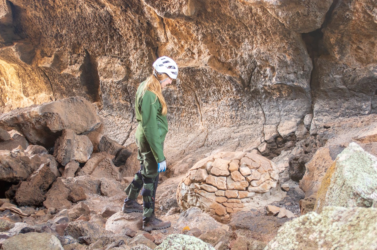 A park ranger dressed in protective caving equipment looks down at a large lava bomb feature on the floor of a lava tube ave.