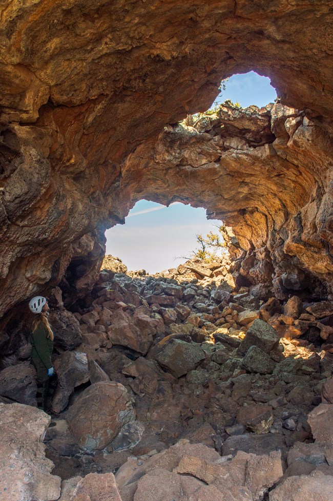 A park ranger in caving gear stands next to an oddly shaped rock at the bottom of a cave with two large openings.