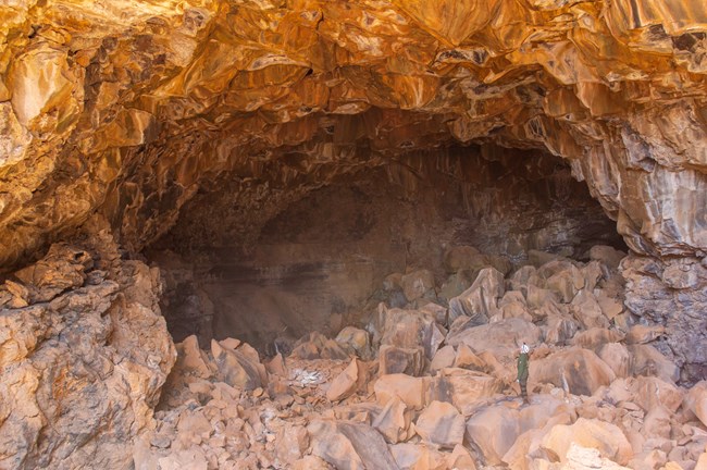 A park ranger in caving gear stands in an enormous cave covered in extremely large boulders.