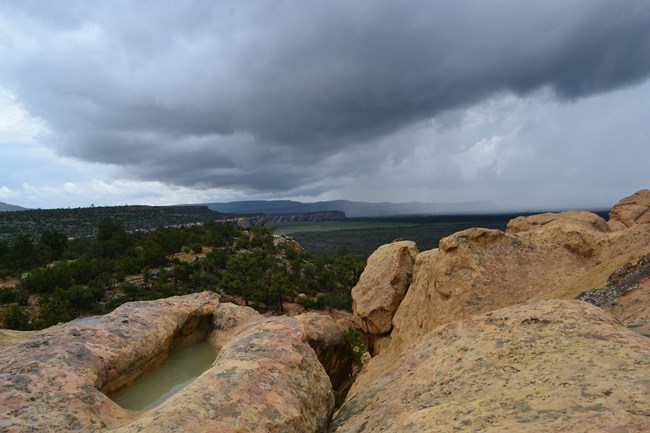 Dark clouds fill the sky above pale yellow sandstone, black lava below, and distant cliffs.