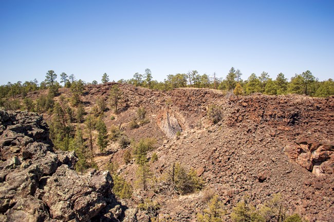 A deep ravine with large boulders and sheer walls of volcanic rock and trees at the bottom and along the rim of the pit.