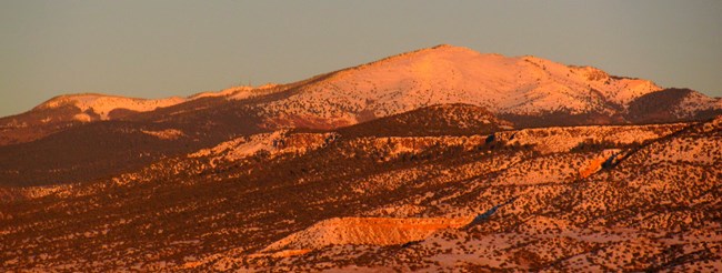 A snow-covered mountain peak and flat-topped ridge glow a deep gold from a sunset's light.