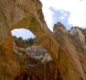 Looking upwards, a light yellow arch with stretches upwards and to the left.  Dark, vertical stripes point up to the clouds above.