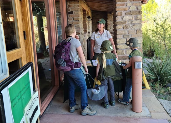 A volunteer stands outside of a building and behind a table, talking to a mother with two young children