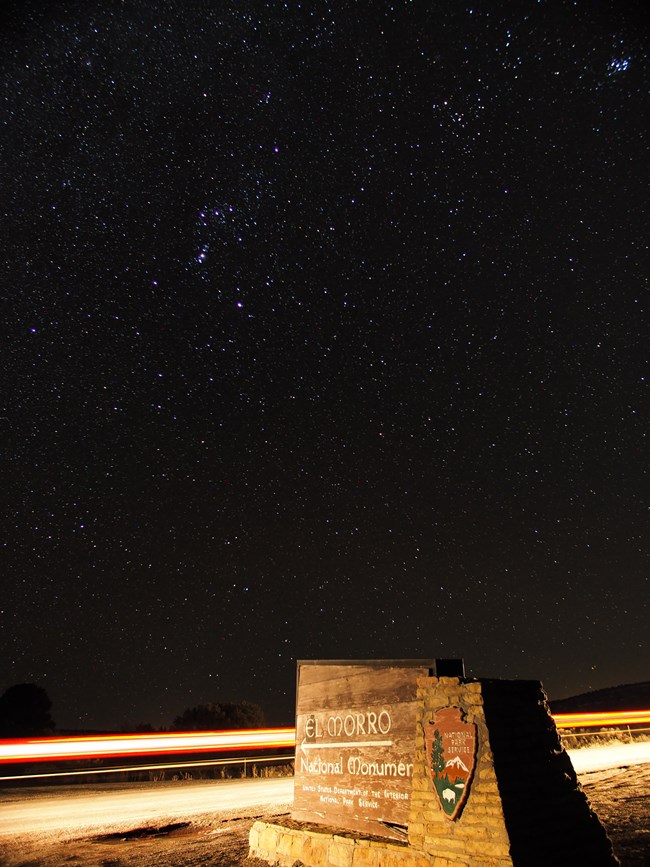 The El Morro National Monument entrance sign at night, illuminated by passing car headlights. Hundreds of stars shine in the sky.