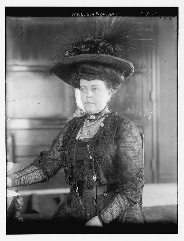 A woman, seated, wearing a black dress and an elaborate black hat