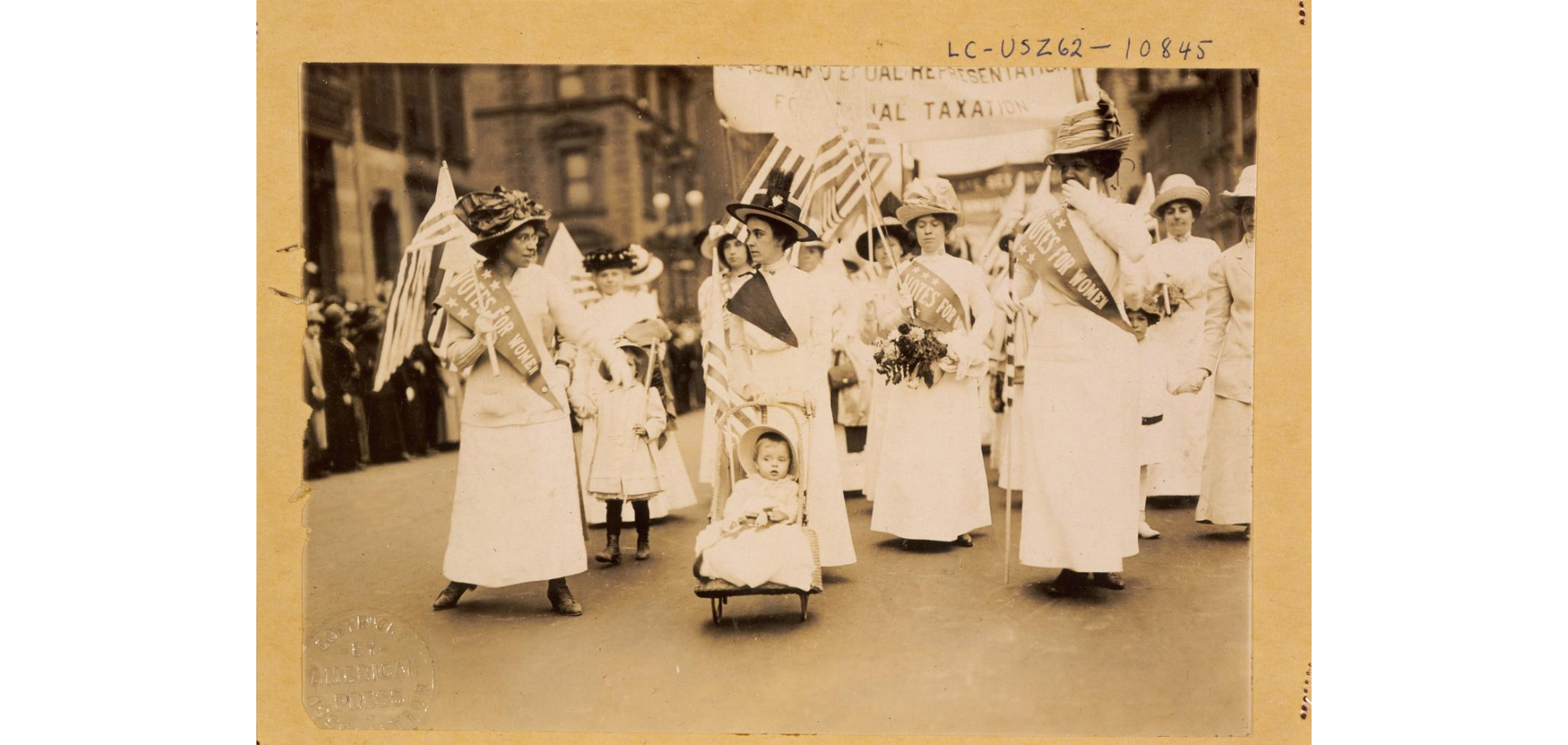 A group of women, wearing sashes and carrying banners, march in a parade to promote women's suffrage. A woman in the center of the group is pushing a baby in a stroller. 