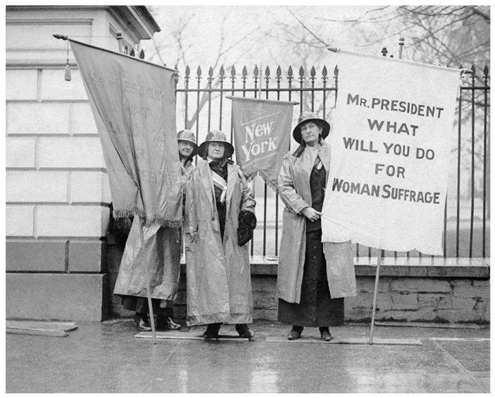 A group of women wearing raincoats standing in front of an iron fence carrying banners with messages of support for women's suffrage.