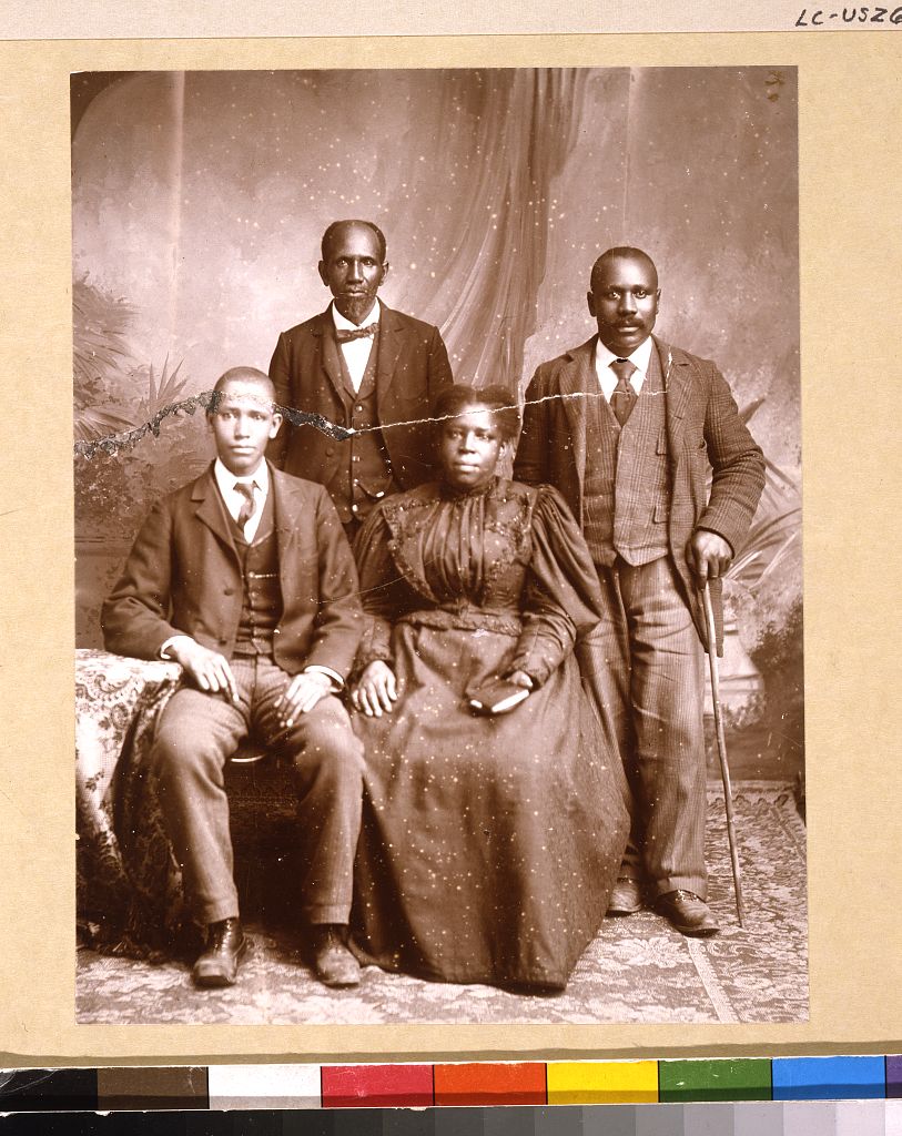 Two men in typical 1890s fashions stand behind a man and a woman, who are seated