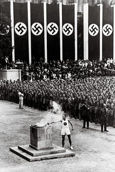 A person in a white athletic suit lights a large torch as spectators watch