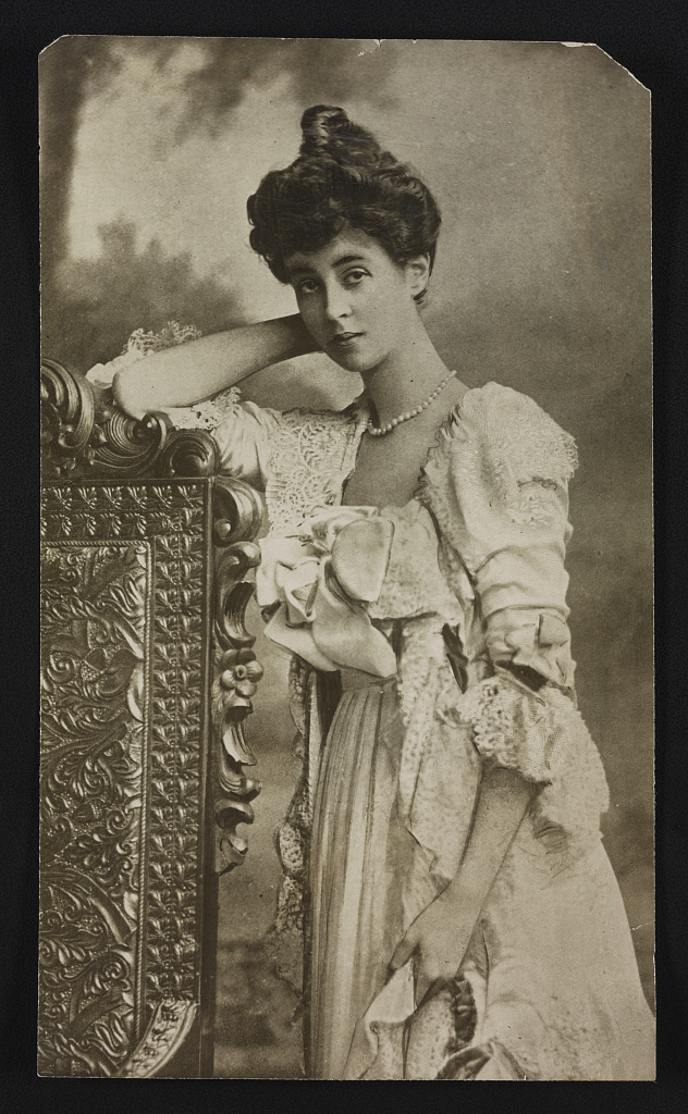 A young woman in an elaborate gown leans on the back of a chair.