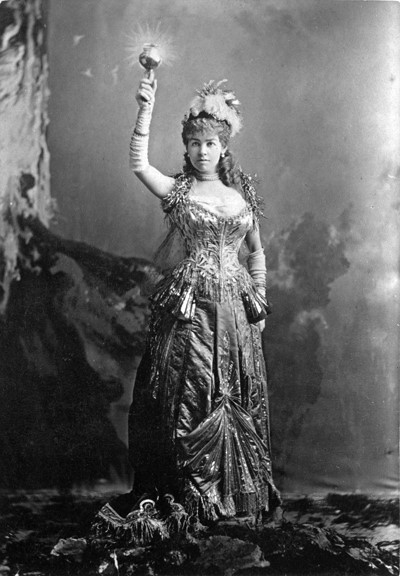 Alice Vanderbilt as an Electric Light A woman dressed in an ornate, metallic ballgown. She is also wearing long gloves and feathered hat. One of her arms is raised and in that hand she is holding a torch.