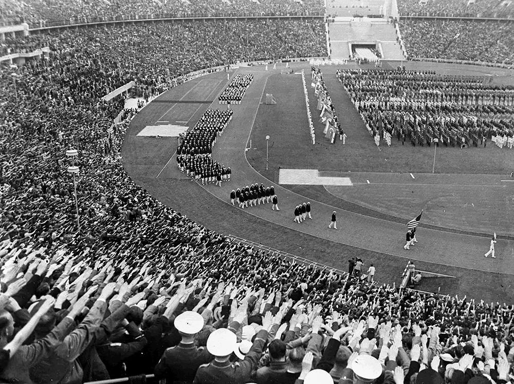 A group of people march along a track, while those in the stands around the track salute them
