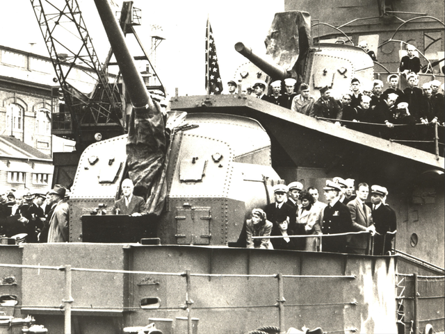 A crowd of people gathers at the front of a military ship to listen to one make speak from a podium