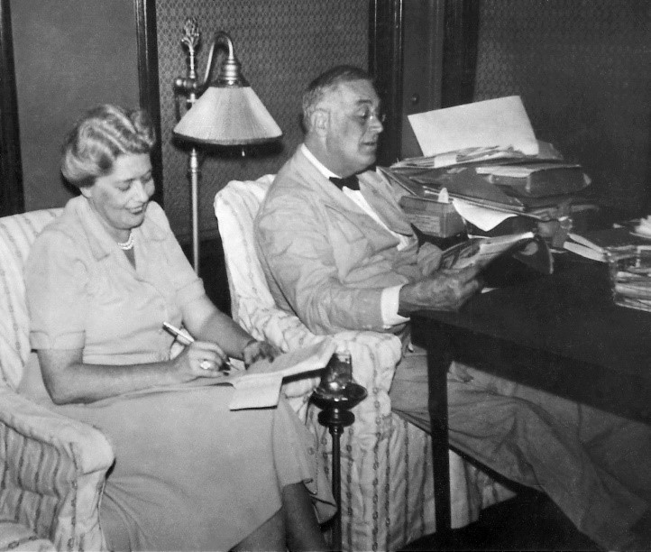 A man, seated, reads a magazine. A woman, seated, to his left, holds a pen and notepad.