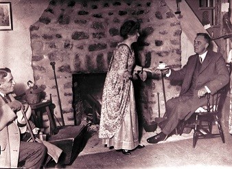 A woman serves a cup of tea to a man sitting by a fireplace.