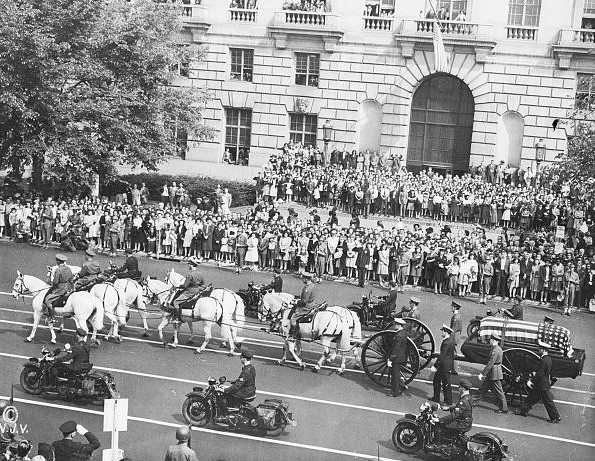 A crowd of people lines a street to watch a horse-drawn carriage containing a coffin go by