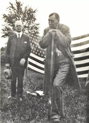 A man leans on a shovel. Another man watches from behind him. There is an American flag in the background.