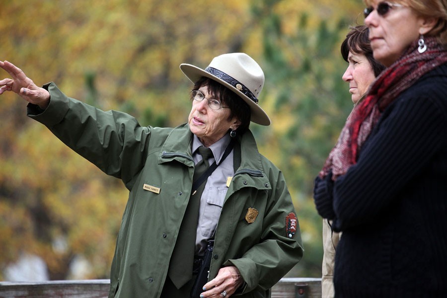 Ranger Marge A woman wearing a Stetson-style hat and a green coat and tie points to something out of frame. Two other women stand to her right.