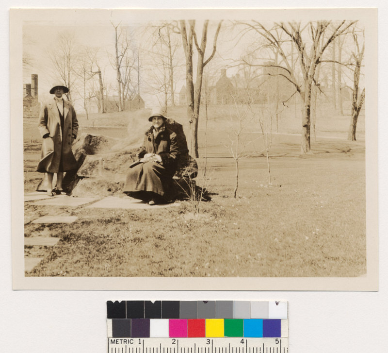 A woman sits on a rock while another woman stands next to the rock. There are trees and buildings in the distance