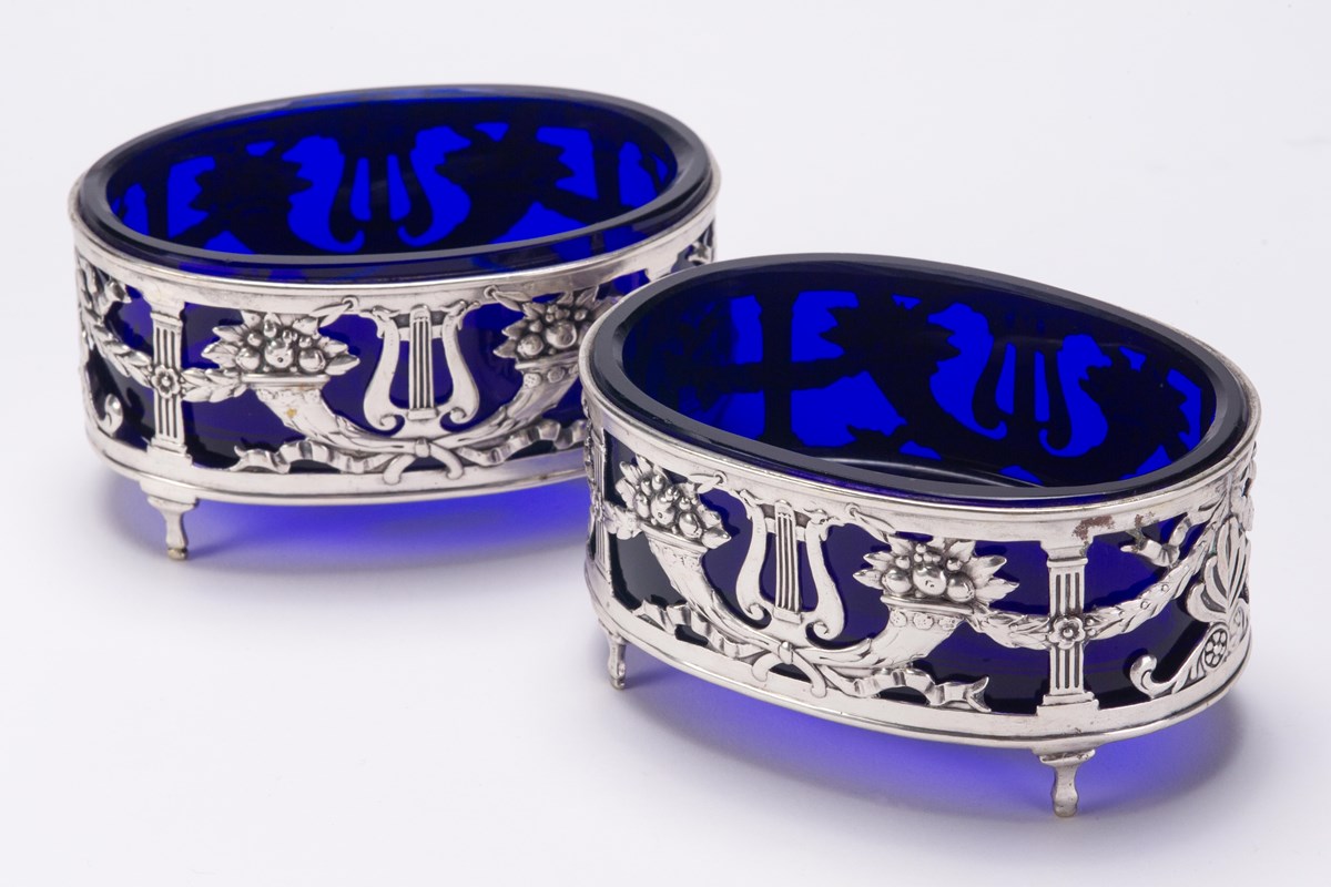 A pair of oval shaped blue glass containers resting in silver holders decorated with cornucopias and lyres.