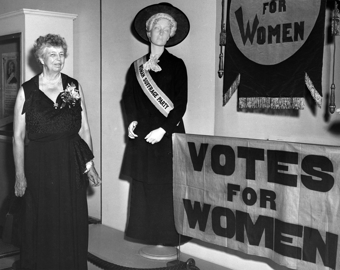 A woman wearing a formal gown stand near an exhibit on women's suffrage.