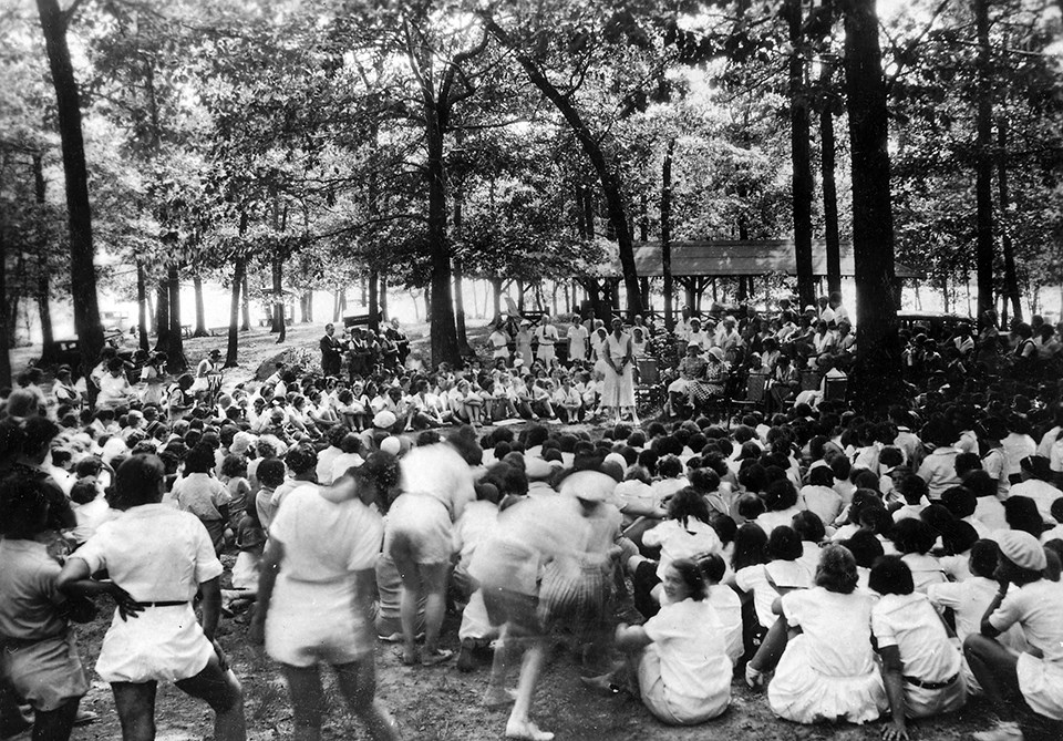 A large group of women gathered around a woman speaking in a forested park.