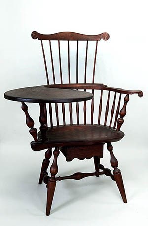 A wood spindle back armchair with writing surface on one arm and a drawer under the seat.