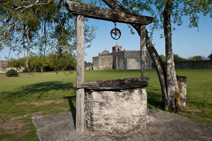 a well in the foreground with a tree hanging over it and a Spanish mission in the background, green grass, blue sky a well in the foreground with a tree hanging over it and a Spanish mission in the background, green grass, blue sky