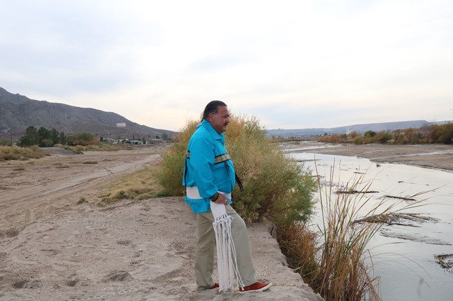 Un hombre indígena con camisa azul y faja blanca está parado en una ribera arenosa de un río manso.