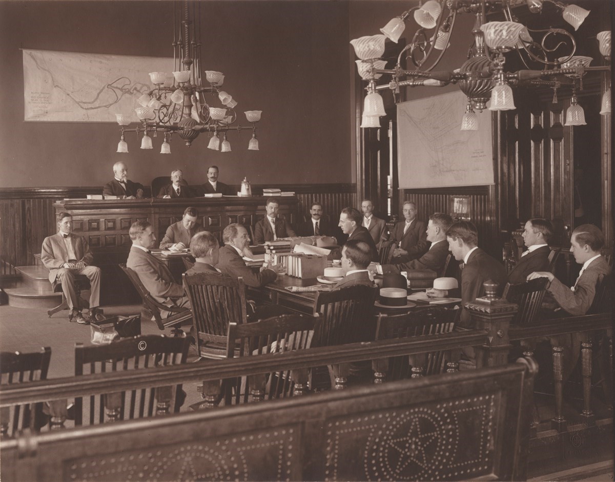 Una foto en blanco y negro de 15 hombres sentados en una mesa de una sala ornamentada y con mapas fijados a las paredes. Tres comisionados están sentados en el estrado elevado en el fondo de la sala.