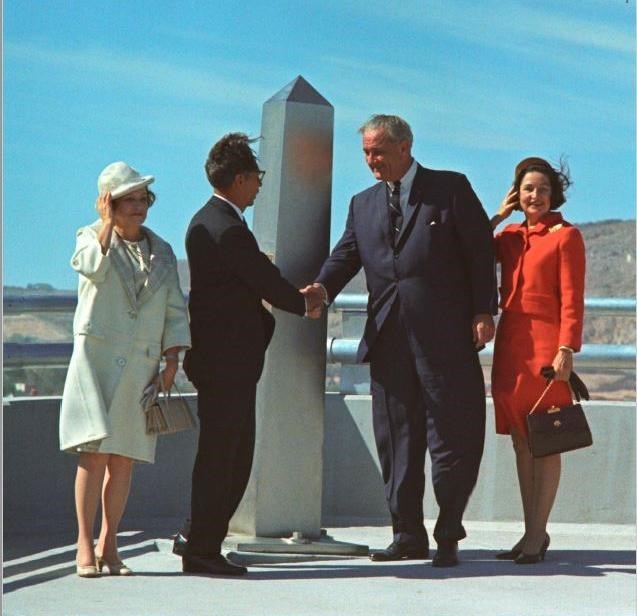 Los presidentes Díaz Ordaz y Johnson dan la mano frente a un monumento internacional en este foto a color. Sus esposas están paradas a sus lados agarrando los sombreros con la mano contra el viento.