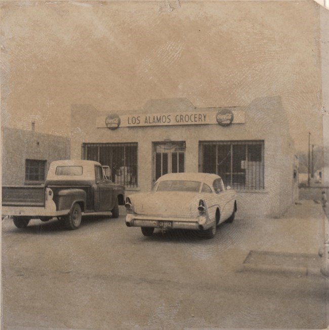 Una foto en blanco y negro de un supermercado de una planta. Una camioneta y un coche de la época de los 1950 están estacionados afuera. Un rótulo sobre la entrada lee en inglés Supermercado Los Álamos y tiene el logotipo de CocaCola a cada lado.