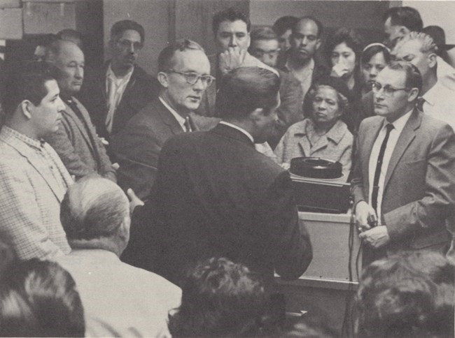 Una foto en blanco y negro de un cuarto lleno de personas reunidas en discusión alrededor de un proyector de diapositiva.