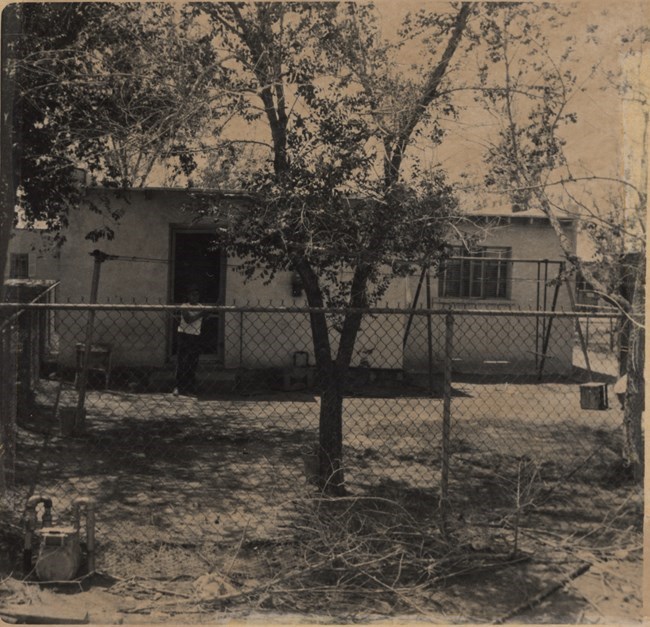 Una foto en blanco y negro del patio de una casa de una planta. Un niño está parado al lado de la puerta dentro del área cercada con maya metálica. Hay un columpio al lado derecho de la casa.