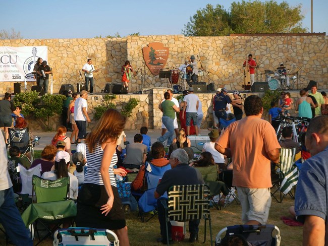 un grupo de personas, unos sentados y otros bailando, visto de atrás mirando hacia el anfiteatro donde toca una banda