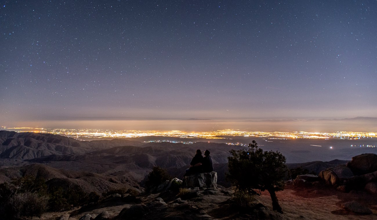 Dos personas sentados en una roca mirando a la ciudad lejana en la noche con el cielo oscuro lleno de estrellas.