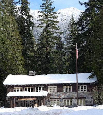 Edificio rústico con su techo cubierto de nieve y enmarcado por árboles coníferos.