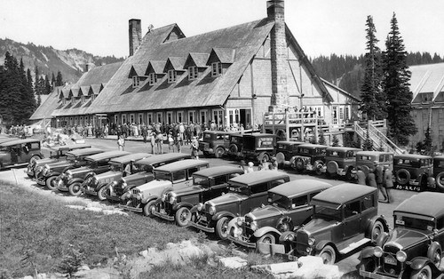 : Una foto histórica en blanco y negro de un gran edificio de madera y piedra con coches antiguos estacionados delante.