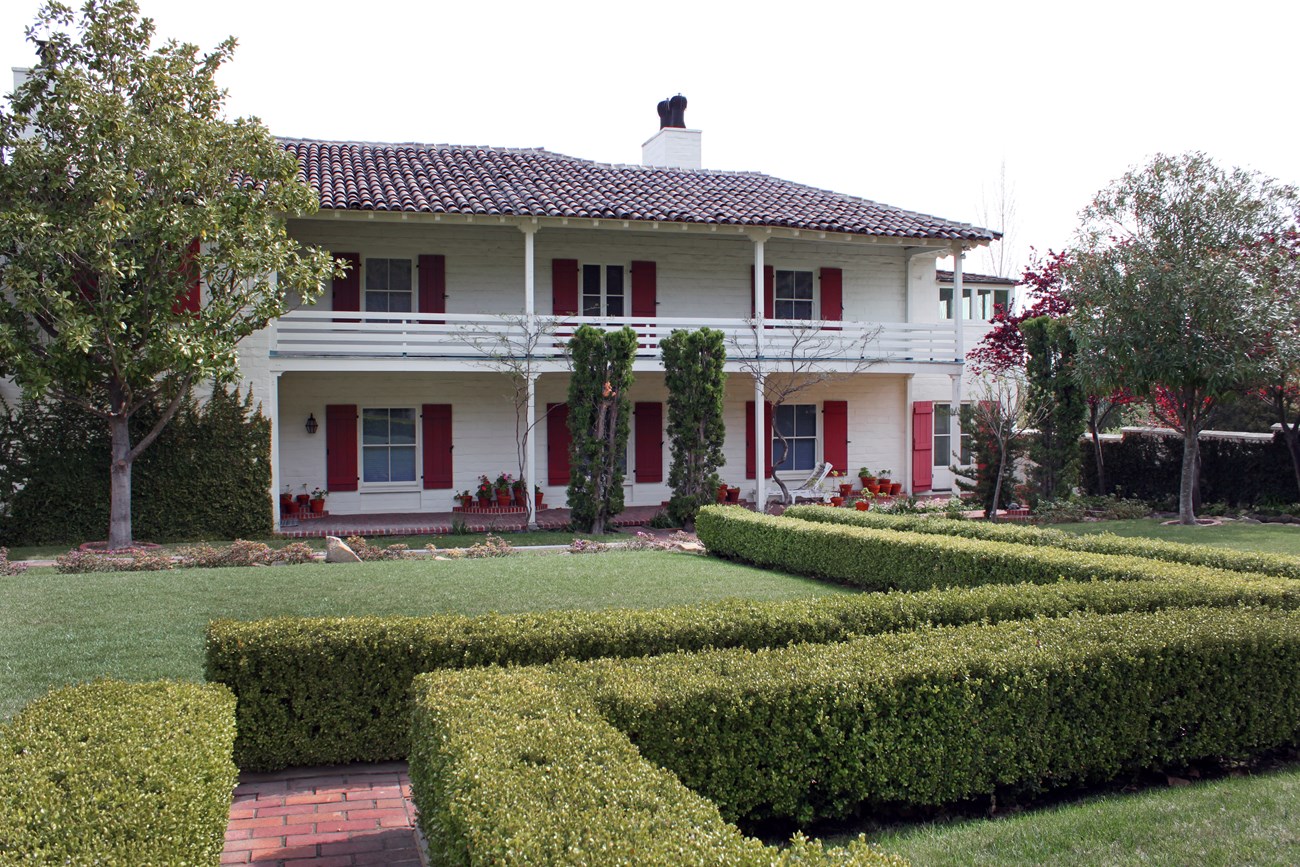 A two-story house with shutters and an upper porch. Plants and bushes are seen along with a brick pathway.