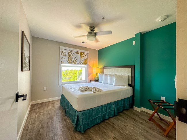 A room with tropical furnishings and colors has a bed, nightstand, ceiling fan and luggage rack. One wall is painted teal and the bed skirt matches.