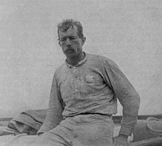 A black and white photo of a man leaning on an object, wearing a long sleeved shirt with a badge