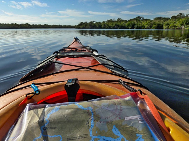 Close up of the bow of an orange kayak. A map can be seen and in the background are water and trees.