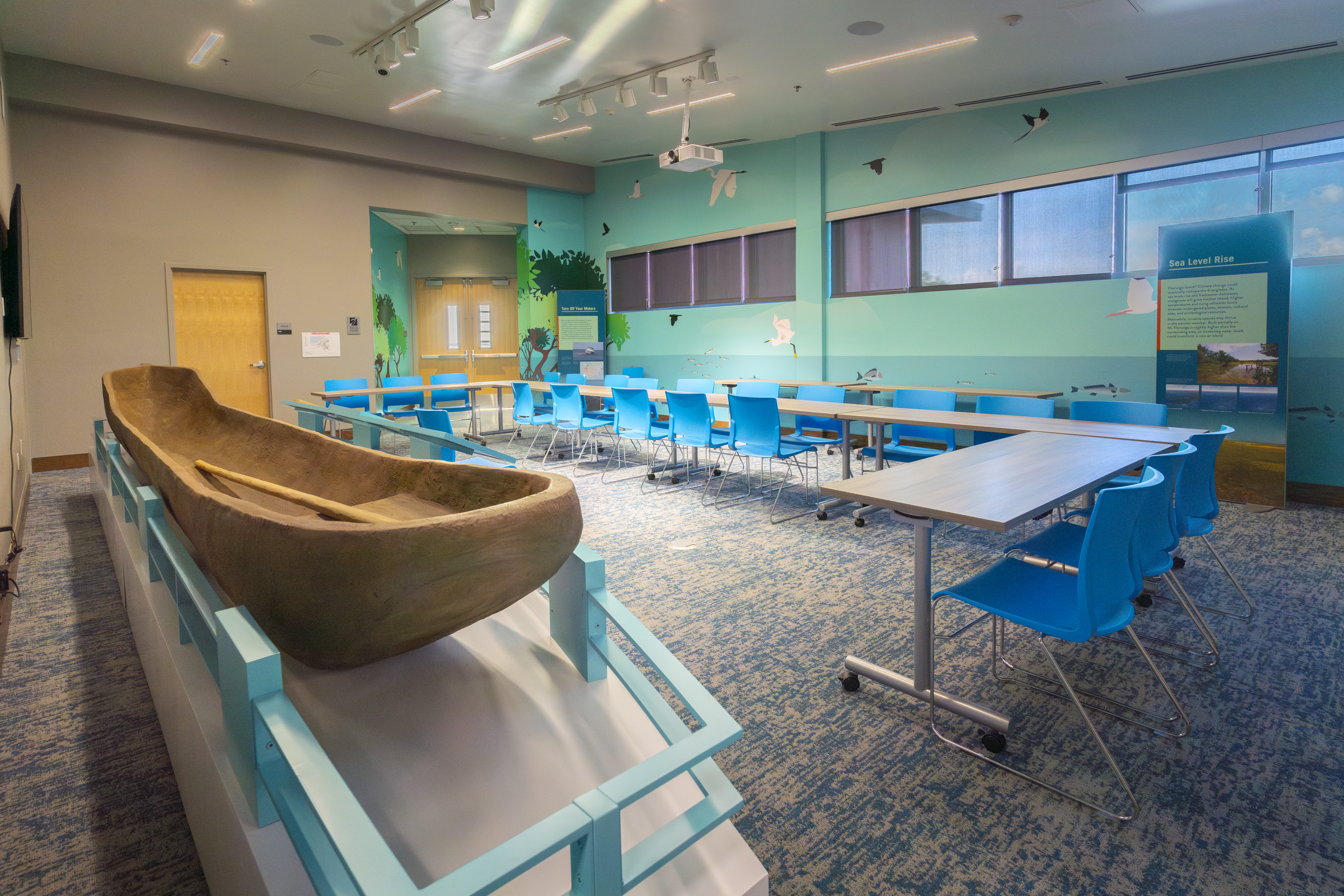 Large tables in a u-shape set up for a meeting in a room with decorative wallpaper and a replica canoe